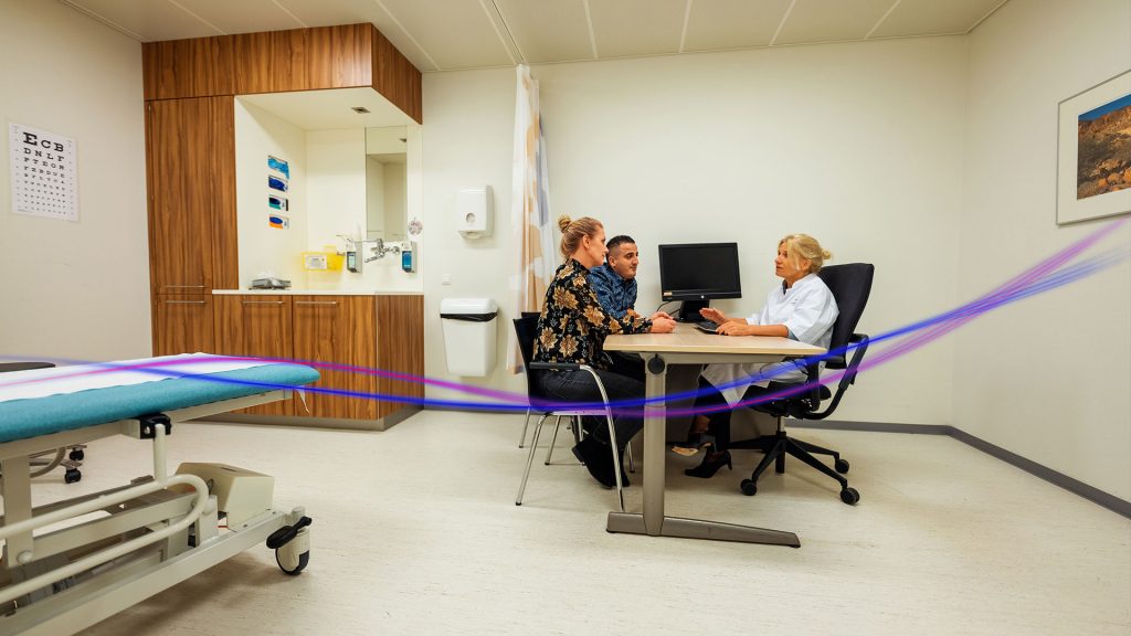 Vrouw en man spreken in behandelkamer met vrouwelijke arts, zittend aan een overlegtafel